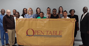 A diverse group of Exact Sciences and Open Table representatives smile while holding a large yellow banner reading “Open Table: Relationship Transforms Communities” against a gray wall.