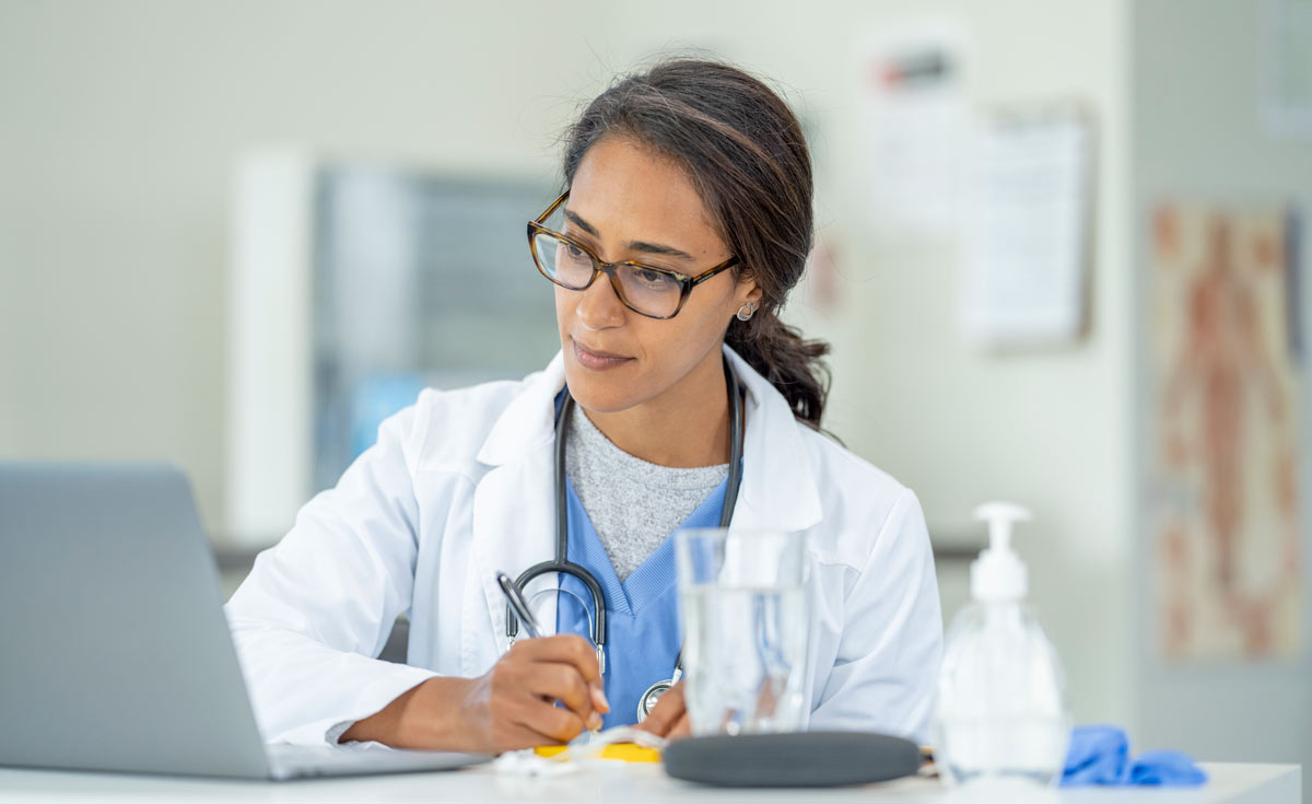Healthcare provider taking notes while looking at a laptop in a clinical setting.